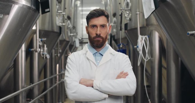Crop view of attentive young male worker looking to camera Handsome man in white lab coat standing at rows of steel brewing vats and crossing hands .Concept of brewery and beer plant