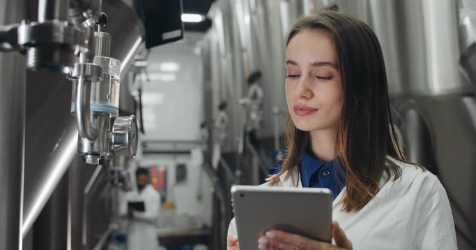 Close up of female brewery worker looking at equipment gauge and entering data in tablet. Woman in white lab coat standing at stainless rows of brewing vats. Male worker at background