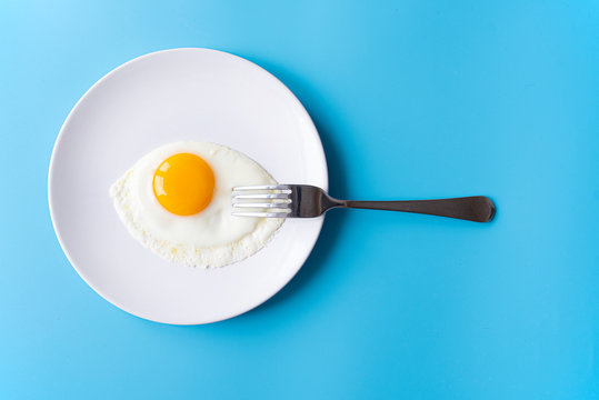 Breakfast, fried egg, yolk, omelet and fork on white plate with color table, creative picture
