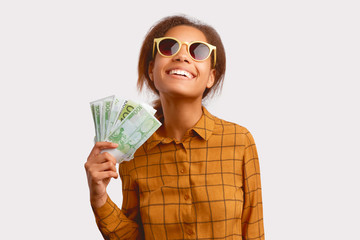 Stylish young woman holds large amount of cash in her hand. Concept of financial success, wealth and prosperity. Portrait of cheerful woman in sunglasses in a checkered shirt isolated on white wall