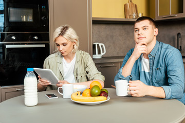 woman using digital tablet near dreamy husband while having breakfast in kitchen
