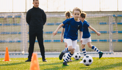 Happy boy practicing soccer on the pitch. Young coach coaching kids football on school sports venue