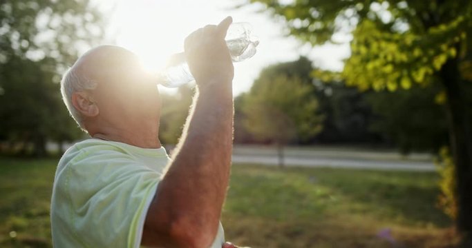Happy elderly sportsman sipping fresh water from bottle while resting during fitness workout in green park