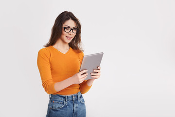 Fototapeta premium A young girl in glasses and a yellow sweater is holding a tablet and smiling. Portrait of a girl on a light background with space for text
