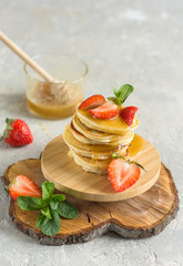 Pancakes with strawberries and honey on a wooden stand decorated with a sprig of mint, a jar of honey in the background