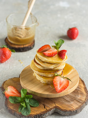Pancakes with strawberries and honey on a wooden stand decorated with a sprig of mint, a jar of honey in the background