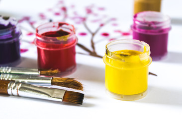 On a white sheet of paper are multi-colored gouache in a jar and brushes for painting. Set for drawing, creativity and hobbies.