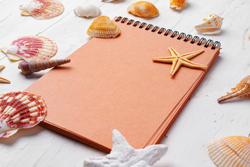 Brown open notepad surrounded by sea shells on white wooden table, copy space