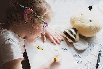 A little girl with glasses draws a portrait of a soft toy bee that lies in front of her. Idea for...