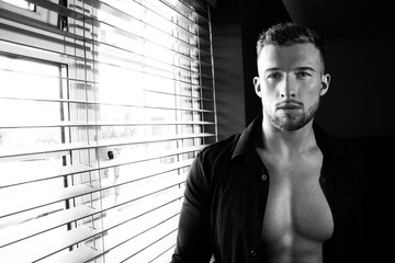 Portrait of handsome man with beard standing next to window with sixpack abs, pecs and unbuttoned...