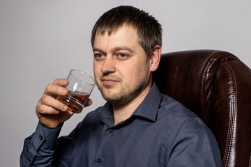 A handsome young man of 30-35 years old in a shirt sits on a brown leather armchair and holds a glass of whiskey in his hand.