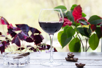 Glass of  wine on wooden table, cigar smoke, pieces of dark chocolate with window green and red plant leaves