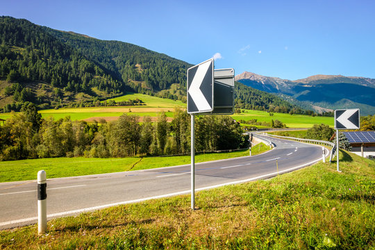 Roadsigns indicates a bend at a bridge over the Rom River in Swiss Val M&uuml;stair, where the sun sets. The road leads to the village M&uuml;stair and then the Italian border into South Tyrol and Val Venosta
