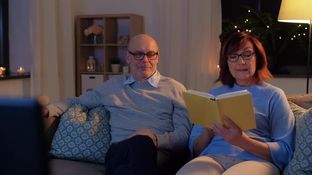 old age, couple and people concept - happy senior man watching tv and woman reading book at home in evening