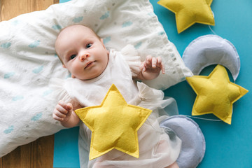 Three month old baby girl in a light elegant dress on a blue background with plush stars and the moon. The concept of baby's restful sleep