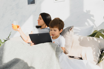 Beautiful smiling mother and son playing with digital tablet laying on bed.