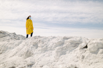 Fototapeta na wymiar Young woman walking on a phosphate mountain that was made by chemical plant waste. She is wearing yellow protective coat and a mask. Ecology protection awareness