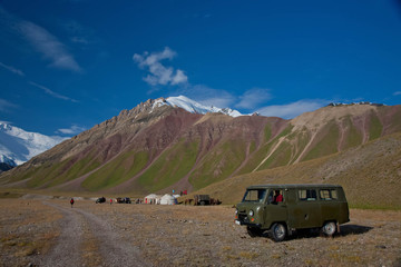 Scenic summer landscape of snowy mountain peaks. Tent camp with yurts in hilly valley. Old car.