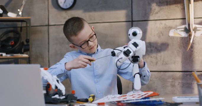 Small nice Caucasian schoolboy putting together or apart a toy robot with a screwdriver while sitting at the table in the school at the technical lesson.