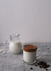 Glass cup with Iced Dalgona Coffee and jug of milk  on grey table. Dalgona coffee is a trend beverage from Korea, it is made from coffee, sugar and hot water. Vertical photo, copy space.