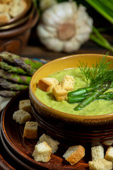 Purred creamy asparagus soup in glass bowl on black plate against raw fresh asparagus and greenery