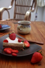 Strawberry cheesecake on light blue plate on wooden platter and wooden table. Rustic mug with milk and strawberry on the side. Bamboo chair in the background. Vertical.