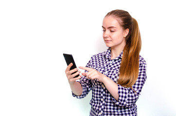 A young girl in a plaid shirt with long hair stands on a white background and holds a smartphone in his hand. A girl looks, is surprised, scared, rejoices looking at a  smartphone
