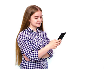 Fototapeta premium A young girl in a plaid shirt with long hair stands on a white background and holds a smartphone in his hand. A girl looks, is surprised, scared, rejoices looking at a smartphone