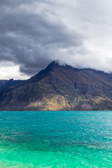 Mountain peaks up to clouds over turquoise water. Rainy day at Lake Wakatipu, New Zealand