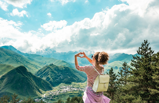 Young female making hands in heart shape sign on the peak of mountains. View in the top of mountain on a hot summer day. Lifestyle concept. Back view. mountain landscape. Freedom concept.