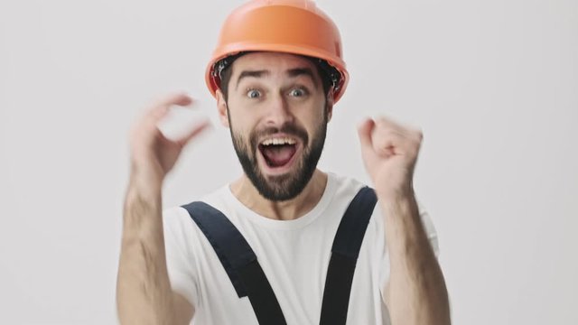 Happy excited young bearded man builder isolated over white wall background in helmet make winner gesture