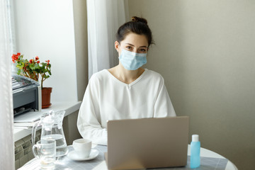 woman in medical mask, hand sanitizer on the table, working with a laptop notebook, online learning education shopping