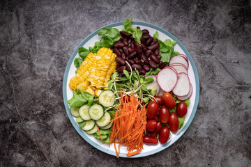 bowl of salad with vegetables ,fresh green oak salad, tomatoes, corn, Red beans, Carrot , red radish, olive oil and cucumber on white marble table. Top view. Food Concept for healthy diet