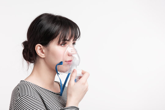 brunette woman holding nebulaizer mask and inhaling a remedy fames into her lungs. side view over gray background isolated picture