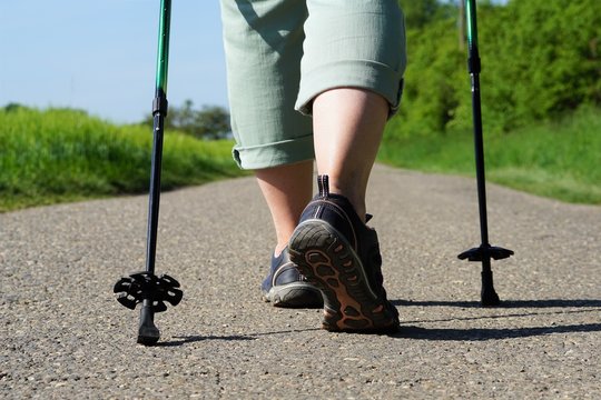 Elderly Woman Does Nordic Walking On An Asphalted Field Road. Female Legs With Nordic Walking Poles On A Cartway At A Sunny Day In Spring.