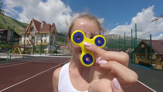close-up game with a spinner: portrait of a happy blonde twirling a yellow spinner in her hand, against the background of a sports field of a summer resort in the mountains. happiness fit activity 4k