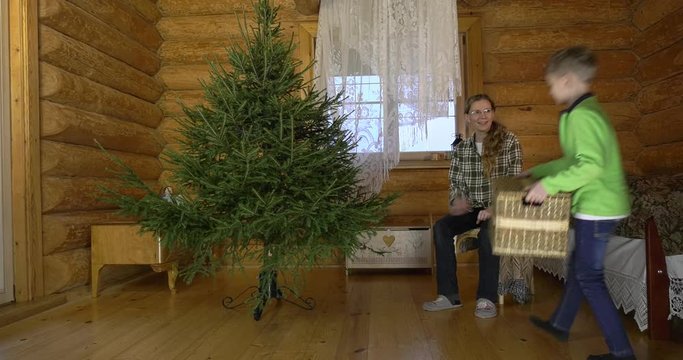Mother with two children dresses up Christmas tree for the holiday. In the interior of the village house, a mother with children amicably take out Christmas toys and hang them on a green fluffy tree.