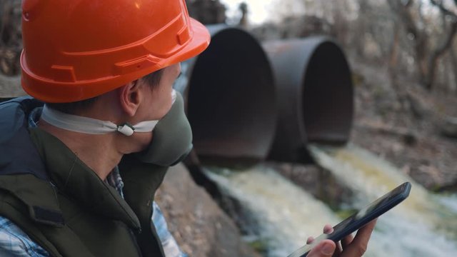 A human ecologist, an engineer in a helmet and a respirator with a tablet measures the level of sewage pollution from the pipe. Environmental pollution.