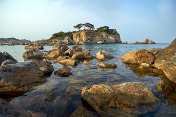 Fototapeta na wymiar Rocky high island in the ocean. Rocky high island in Orlik Bay in the Sea of Japan. In the foreground are stones and clear water. Far East.