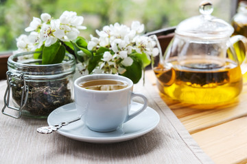 a transparent teapot, a white Cup on a saucer, and loose tea in a jar on a light wooden table. Near the white flowers of Apple or plum trees