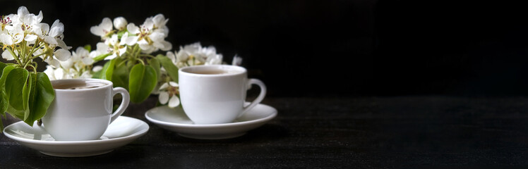 two white cups on a saucer on a dark wooden table. Near the white flowers of Apple or plum trees. long banner with copyspace
