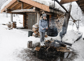 a girl in a blue jacket warms her hands over a bonfire where a cauldron is cooked against a wooden...