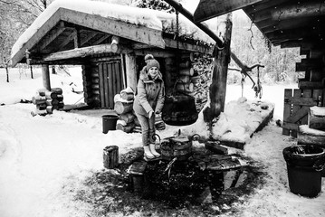 a girl in a blue jacket warms her hands over a bonfire where a cauldron is cooked against a wooden hut. Karelia winter
