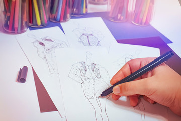 Fashion designer at work. On the table is a white paper with sketches of models with a black pen. He makes drawings of dresses on girls with his hand. In the background are glasses with pencils.
