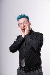 Young stylish european man with blue hair expressing amazement, shock and excitement, opening his mouth widely and touching his face