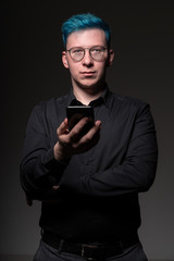 Stylish and unusual man with blue hair in a black shirt is holding is smartphone and looking straight in camera with calm confidence, studio photo