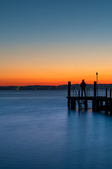 Fototapeta na wymiar A man silhouette standing on wooden pier lonely at the sea with beautiful sunset. lsunset seascape at a wooden jetty.