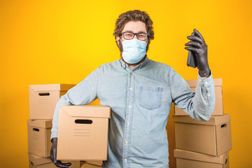 Fototapeta na wymiar Man courier in a medical mask holds a phone with orders in his hands, standing on a yellow background among the parcels