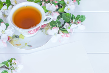 Obraz na płótnie Canvas a cup of tea, a piece of paper and white flowers on a white background. a cup of tea and branches of a blooming apple tree close-up. festive background with a cup of tea and white flowers.