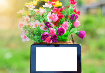 Tablet placed beside the basket containing colorful flowers, with the sunlight shining in the background. Copy space. Selective focus. Flare.